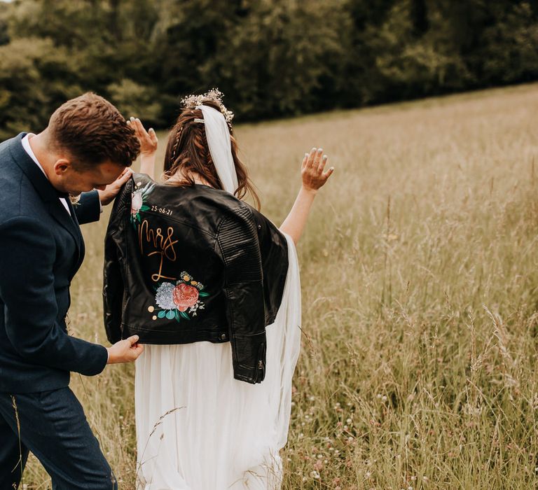 Groom in blue suit looks at brides hand painted bridal leather jacket for the first time in field at garden wedding reception 