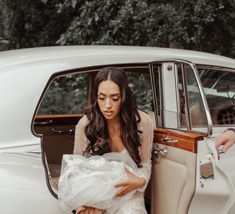 Bride climbs out of white wedding car as she holds her wedding gown in her hand