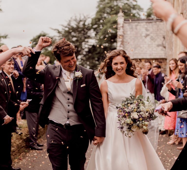 Bride in white Elbeth Gillis wedding gown holding white and green bridal bouquet smiles whilst holding hands with grooming dark brown three piece suit as guests throw confetti outside church