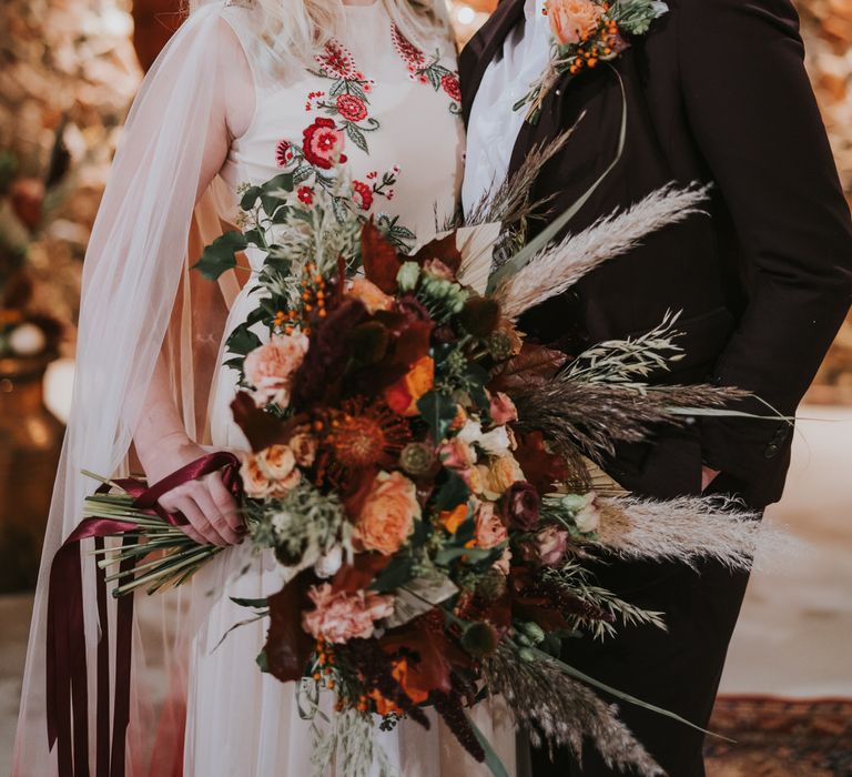 Two brides looking into each other's eyes, one is holding a huge bouquet of assorted orange and red flowers with wild grasses and dried palm leaves