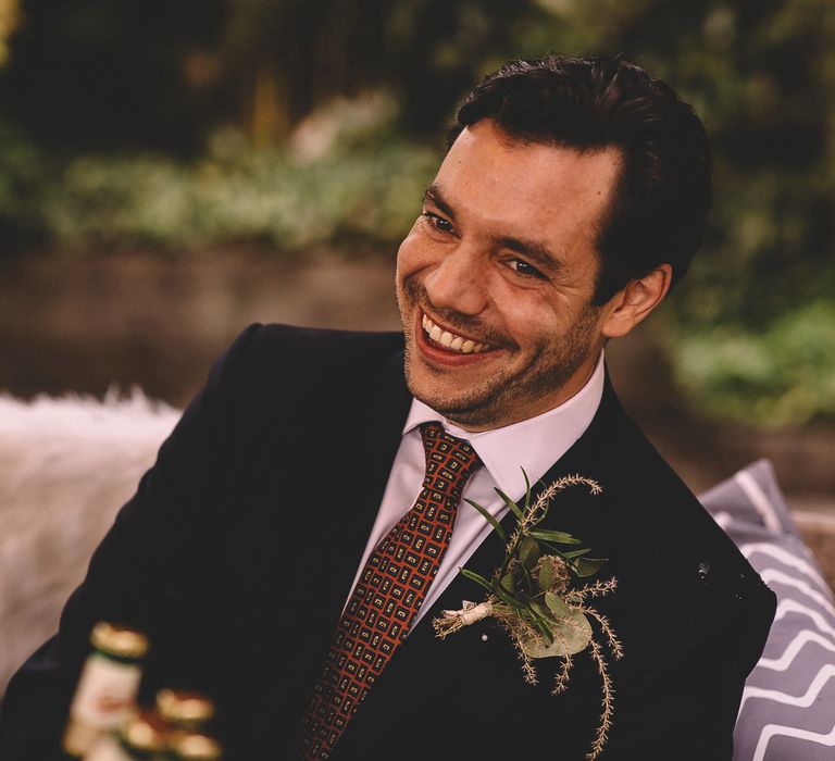 Groom in a navy suit, white shirt and red patterned tie with a green foliage button holding laughing during the pub wedding reception speeches