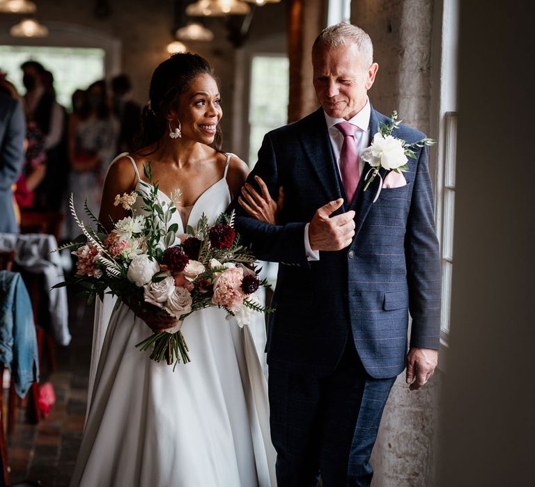 Emotional bride in white cami wedding dress holding white, pink and green bridal bouquet smiles at man in checked suit and pink silk tie as they walk arm in arm down the aisle during wedding ceremony at The West Mill Derby