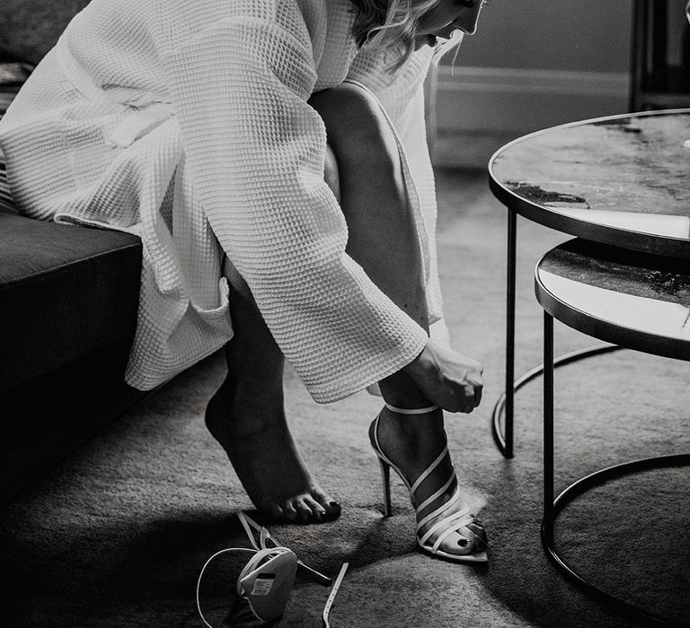 Black and white portrait of the bride on her wedding morning in a waffle getting ready robe putting on her strappy wedding shoes 