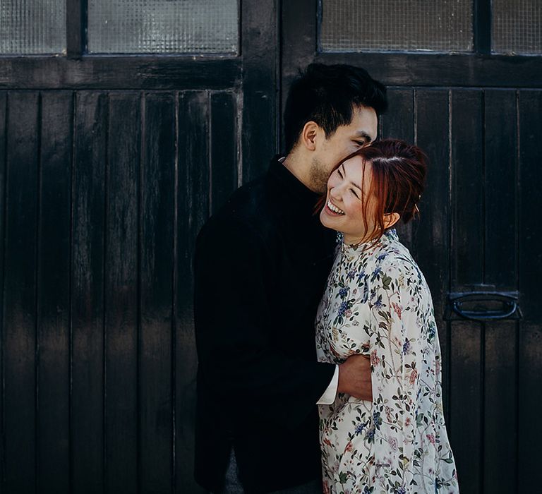 Groom in a velvet jacket kissing his brides cheek as she laughs in a blue floral dress with cape sleeves 