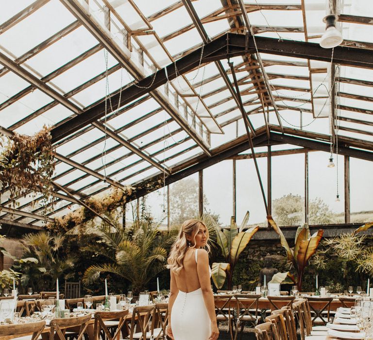 Bride in white open back Made With Love wedding dress stands in glasshouse at Anran Devon for wedding breakfast with long wooden tables, wooden chairs, candles and large fern plants 