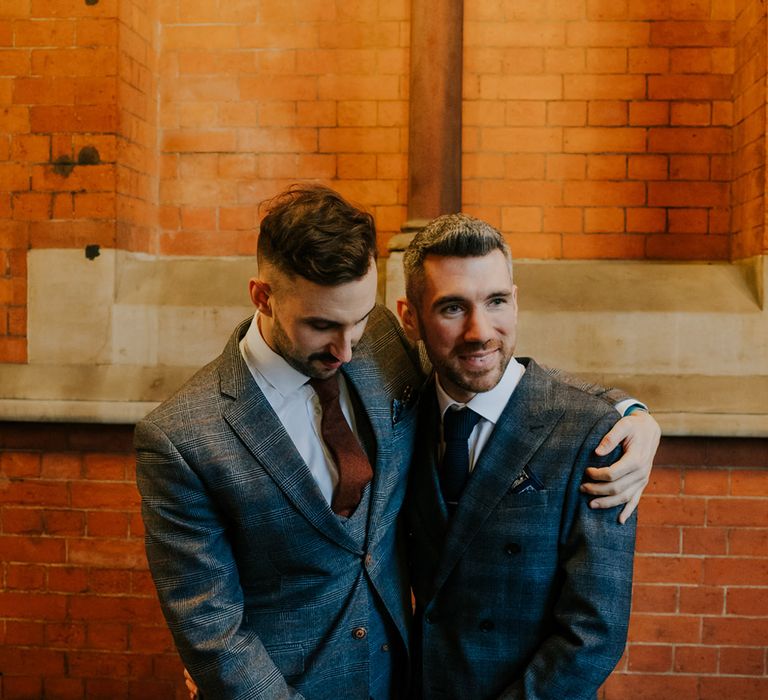 Groom holds his arm around groom as they hold hands in front of brick wall on their wedding day