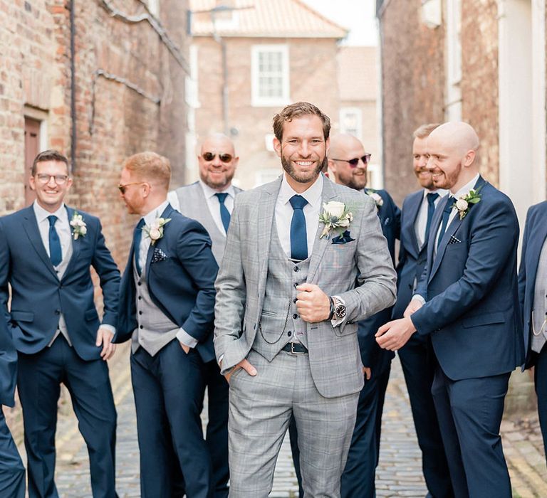 Groom in a light grey check wedding suit with his groomsmen in navy three-piece suits standing behind him