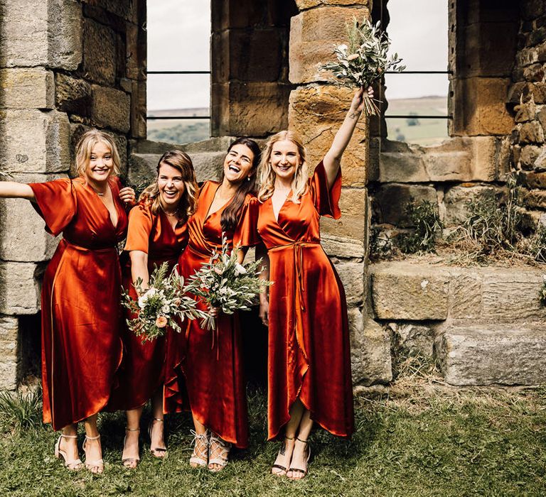 Bridesmaids in wrap deep orange satin bridesmaid dresses by Rewritten waving their green foliage bouquets in the air 