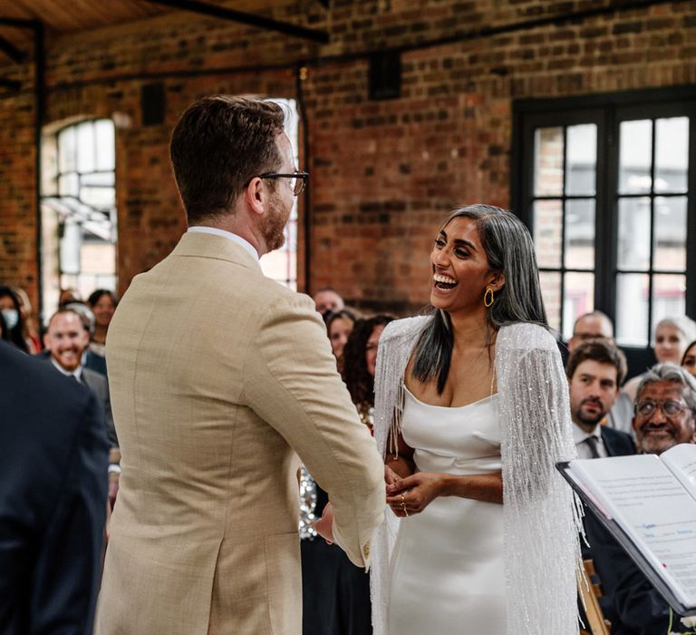 Groom in linen suits stands at the end of the aisle with bride in satin Halfpenny London wedding dress and tasselled bridal cape during wedding ceremony at Loft Studios London