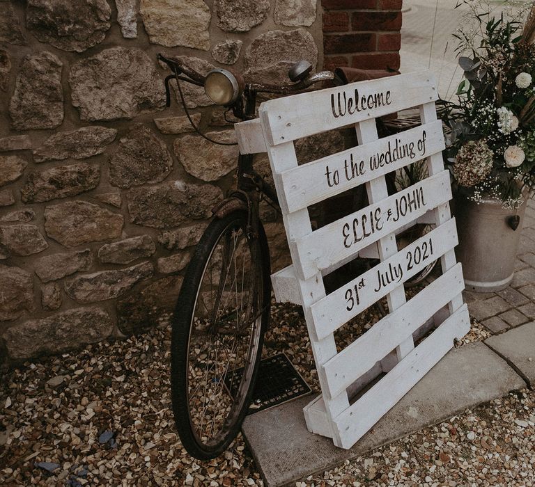 White wooden pallet wedding welcome sign leaning on bike next to milk churn full of florals for Isle of Wight wedding with macrame wedding decor