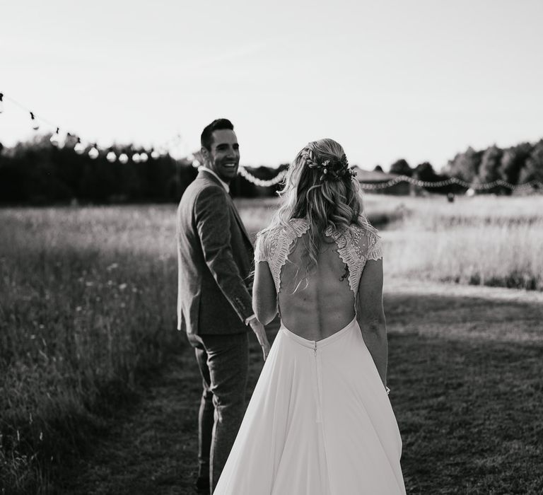 Bride in lace cap sleeved open back wedding dress with train and flowers in her hair walks with groom through field during late summer wedding at Wellington Wood Norfolk