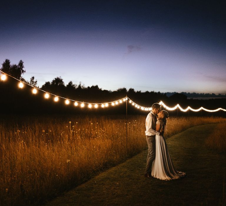 Bride in lace top wedding dress with train hugs groom in white shirt with grey trousers stand in field with festoon lighting during late summer wedding at Wellington Wood Norfolk