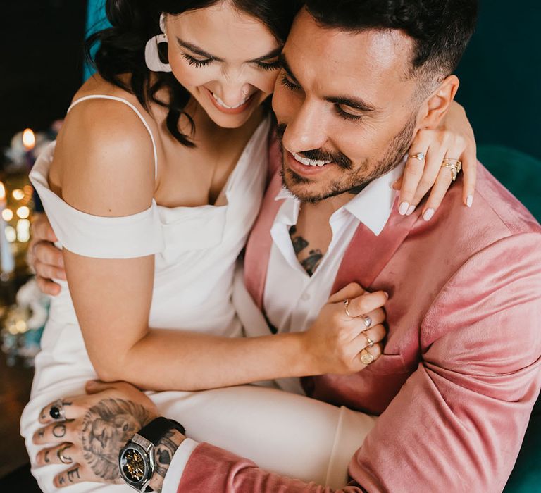 Bride with white nail polish and ring stacks embracing her tattooed groom in a pink velvet jacket 