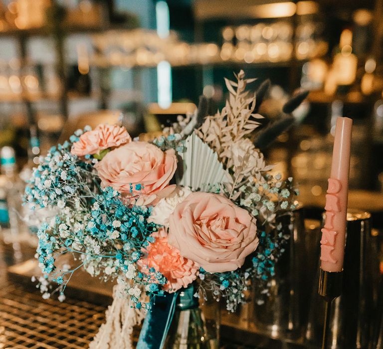 Pink rose and blue gypsophila wedding bouquet tied with teal velvet ribbon resting on the bar 