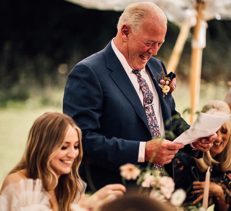 Father of the bride in blue suit and patterned ties makes speech during marquee wedding reception in Cornwall