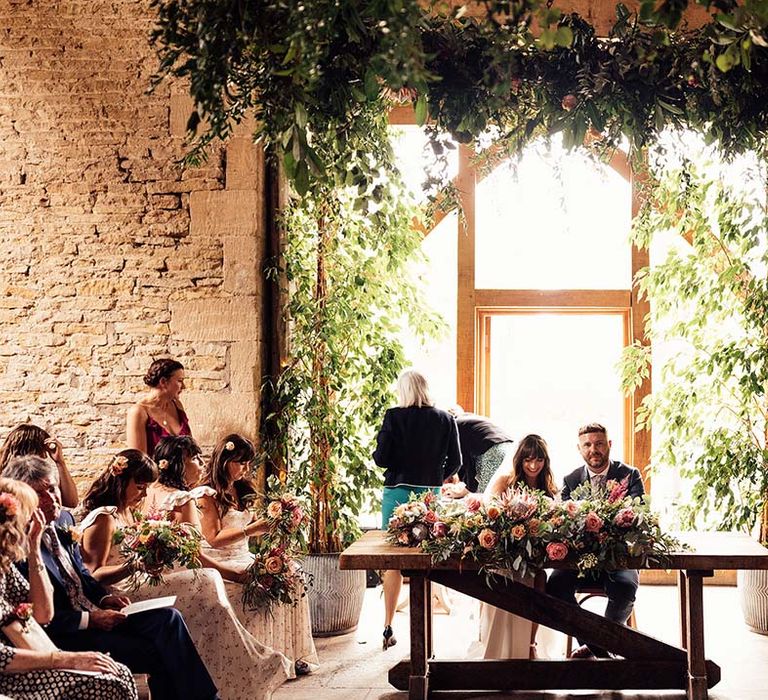 Bride & groom sit in front of large barn window during wedding ceremony as they sign their marriage certificate 