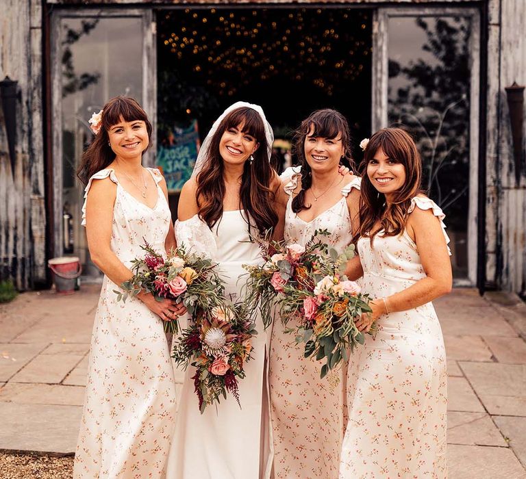 Bride stands with her bridesmaids who hold colourful floral bouquets as floral gowns