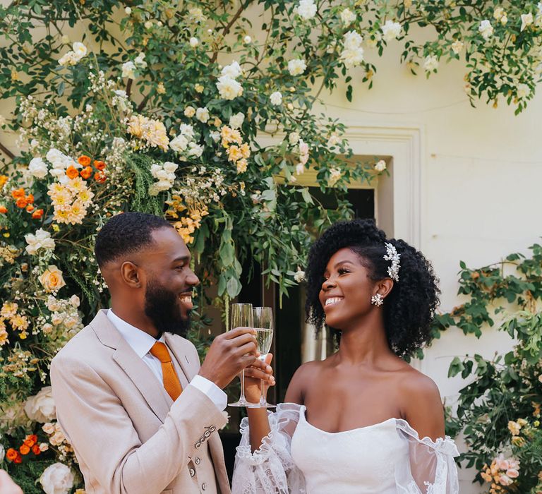 Black bride and groom in a beige suit and strapless wedding dress with sheer sleeves toasting champagne at their outdoor wedding reception 