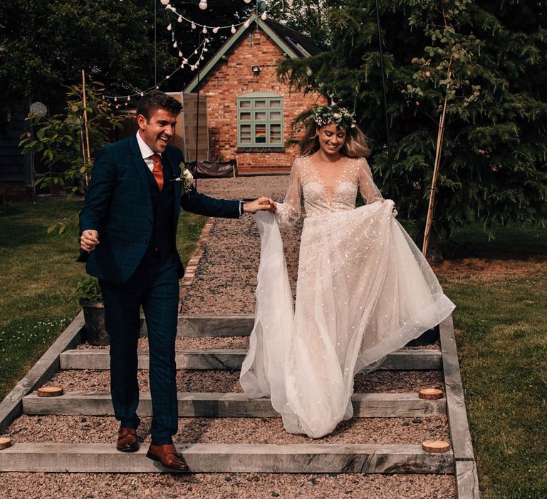 Bride in long sleeve v neck wedding dress and flower crown runs down garden stairs holding groom in blue checked three piece suit at garden wedding reception