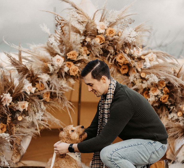 Groom-to-be in jeans, dessert boots, jumper and scarf at boho marriage proposal putting on his pet cockapoo's lead 