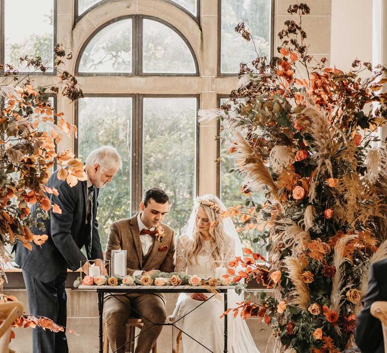 Bride in lace boho wedding outfit and veil sits with groom in brown tweed suit as they sigh wedding registry at intimate wedding ceremony with orange and green floral columns