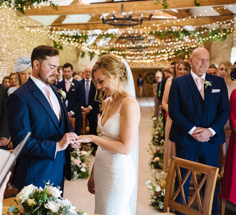 Groom in a navy suit with pink tie putting a ring on his brides fingers during the wedding ceremony 