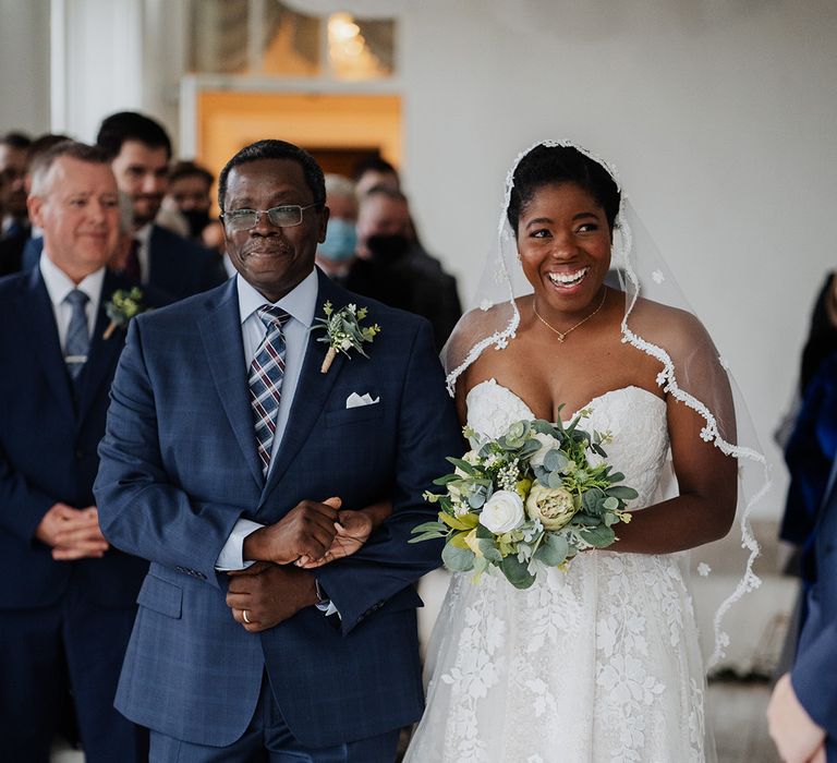 Father of the bride holds daughters hand as they walk down the aisle of her wedding day smiling 