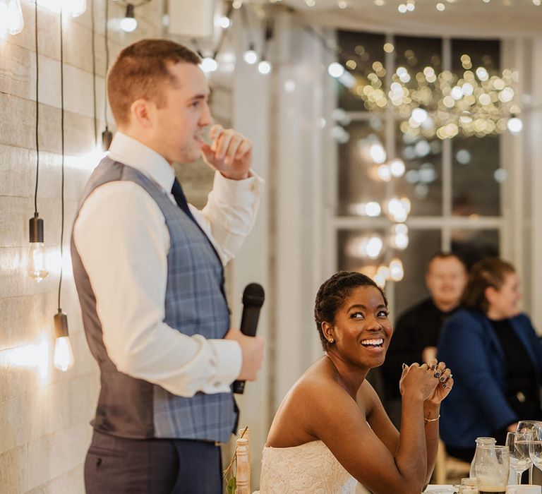 Groom in white shirt and checked waist coat stands to do speech in fairy light reception 