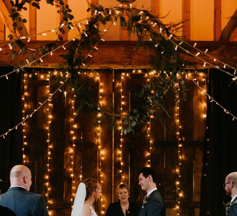 Bride in lace and tulle wedding dress and veil stands in front of fairy light wall with groom in dark suit for rustic wedding ceremony