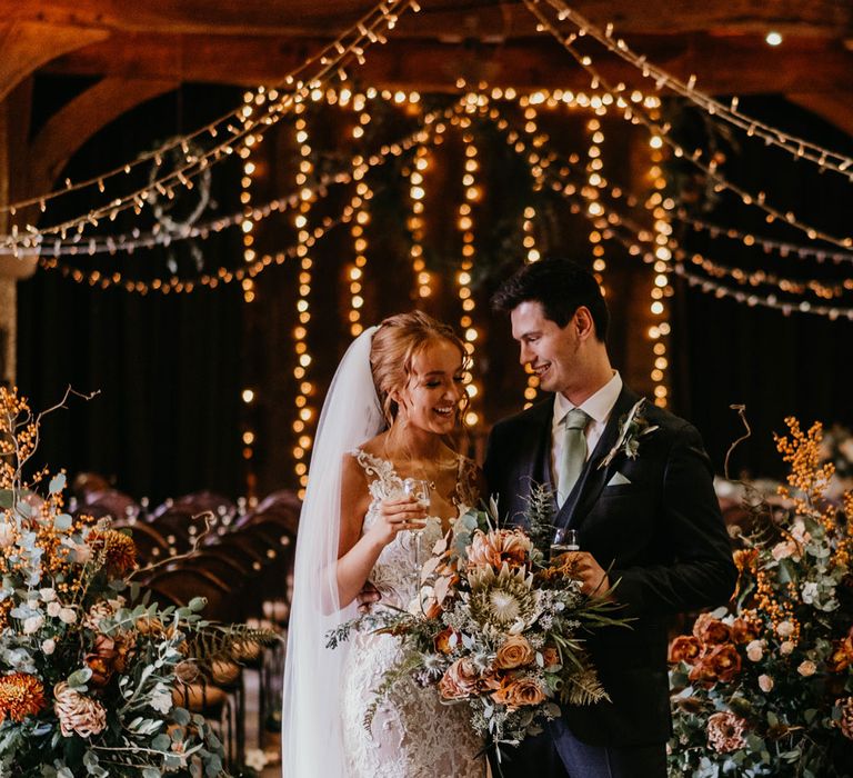 Bride in lace strap wedding dress with tulle skirt and veil holding large orange and green bridal bouquet stands holding a glass of sparkling wine with groom in dark suit and blue tie after rustic wedding ceremony