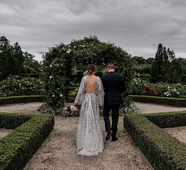 Bride in a low back silver sparkly wedding dress and groom in a navy suit arm in arm in their garden wedding 