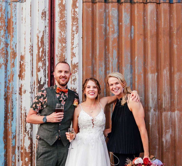 Bride in lace wedding dress holding glass of sparkling wine stands with groom in waistcoat holding pint of ale and wedding guests holding colourful rustic bridal bouquet