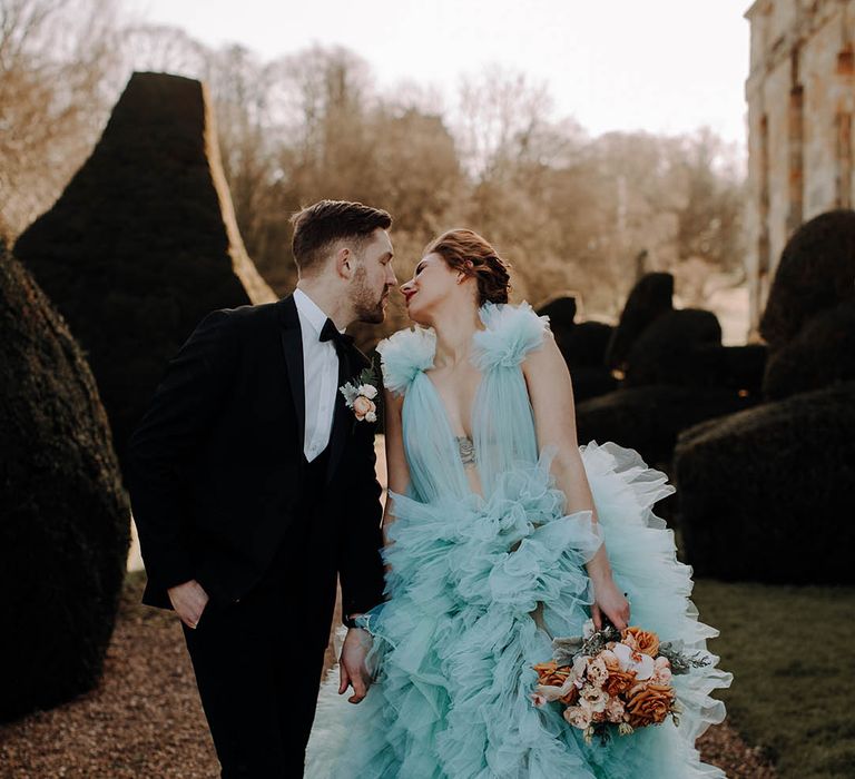 Bride in a blue ruffle wedding dress with plunging neckline kissing her groom in a tuxedo during golden hour 