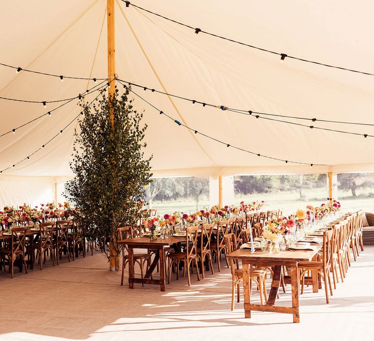 Interior of marquee garden wedding with long wooden tables, colourful florals and black bulb string lights 