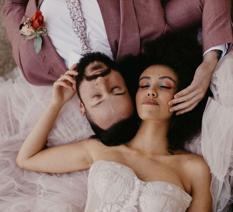 Black bride with afro hair and green eyeshadow laying down with her groom in a pink suit and floral tie 