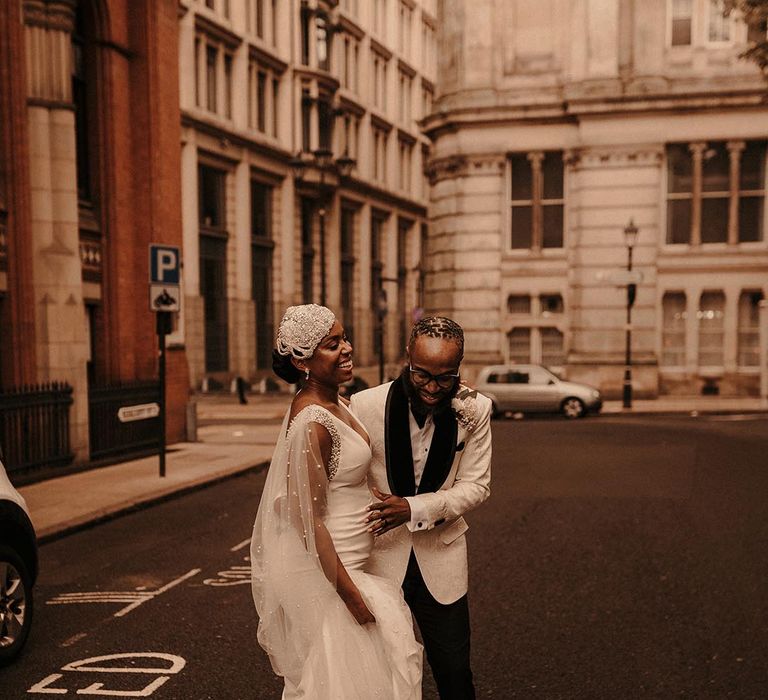 Groom in a white tuxedo jacket dancing with his bride in a fitted wedding dress with Watteau train and an embellished cap headdress in the streets of Birmingham 