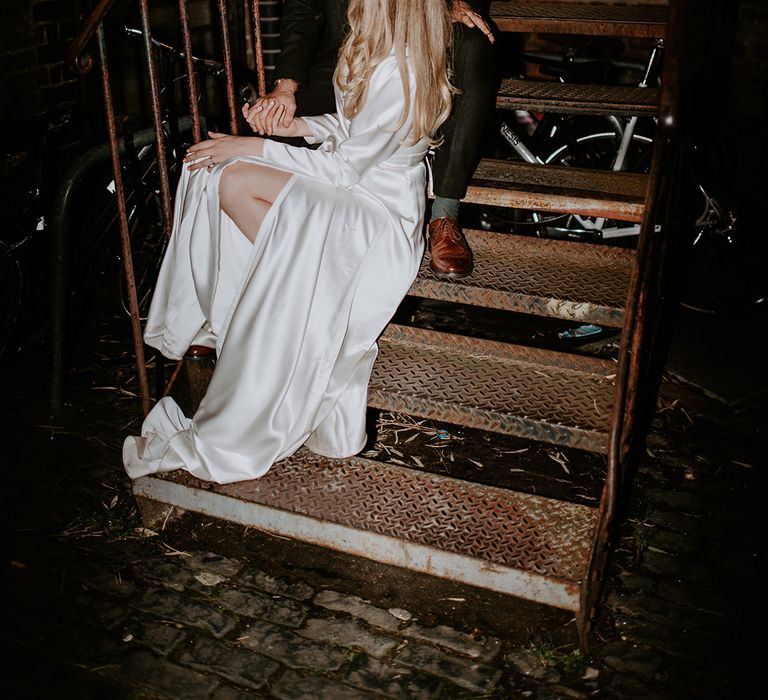 Bride & groom sit on staircase and smile lovingly at one another