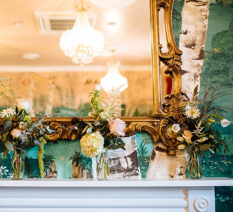 White fireplace with small vases of flowers in front of golden mirror on forest themed green wallpaper 