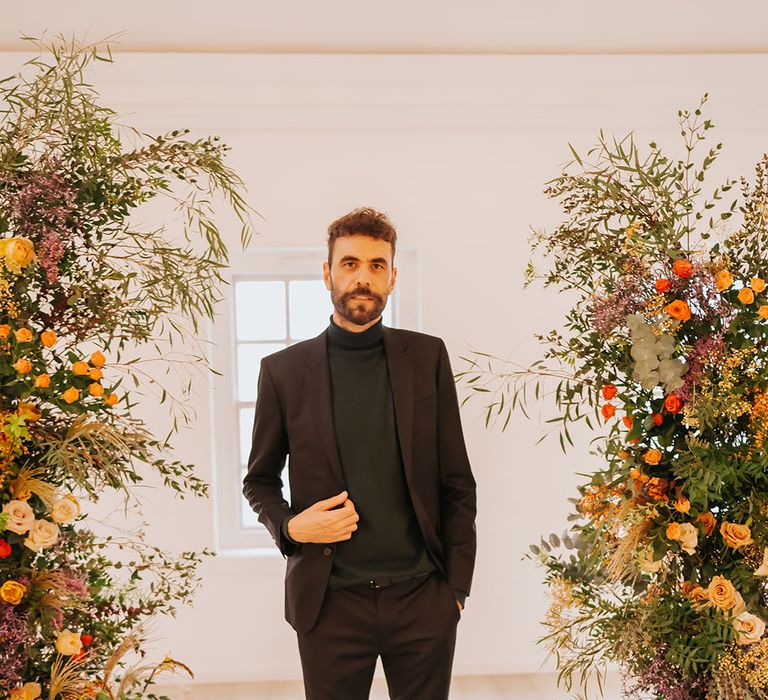 Groom in an all-black suit and top standing in-between orange, yellow and foliage column wedding flower arrangement 