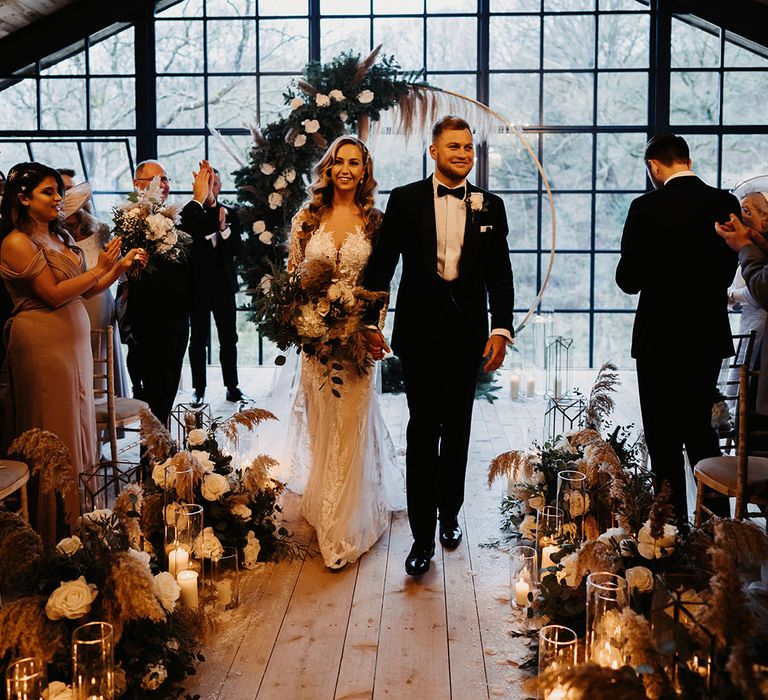 Bride in lace wedding dress and groom in black tie walk back down the aisle as a married couple with floral moongate at the altar