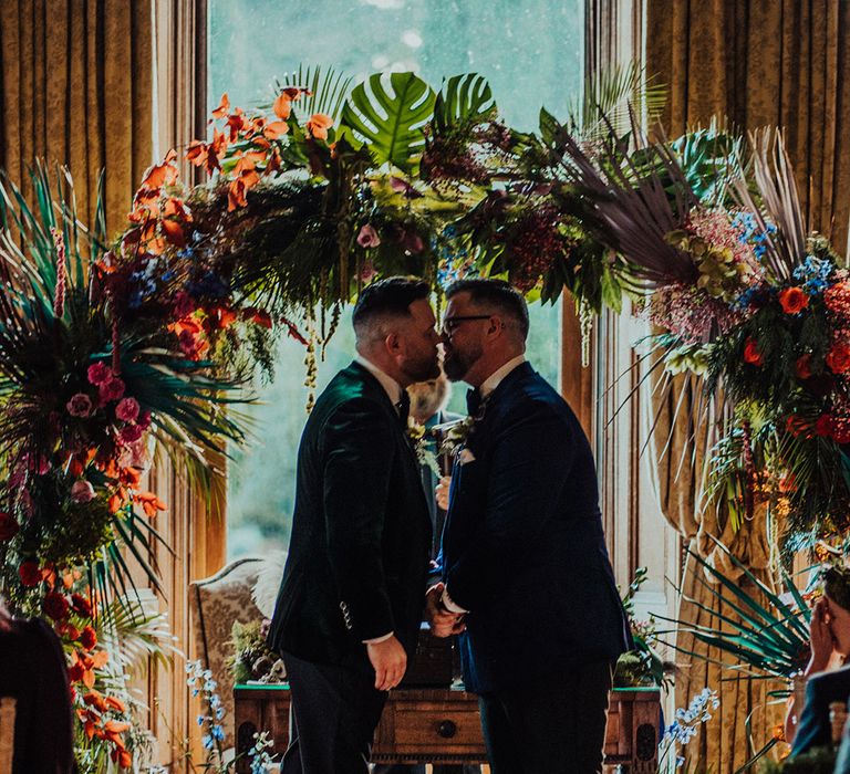 Grooms in blue and green velvet tuxedo jackets share their first kiss as a married couple in front of tropical flower moongate