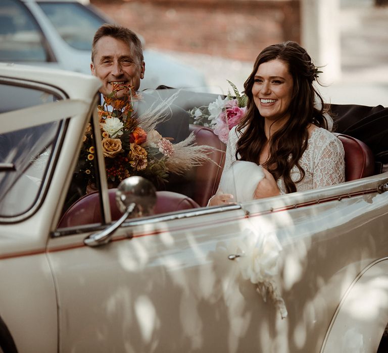 Bride in lace wedding dress sits with father in white vintage wedding car transportation 