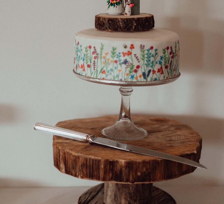 Wedding cake with white frosting with colourful flowers and bees with custom wooden cake toppers of bride, groom and their cat