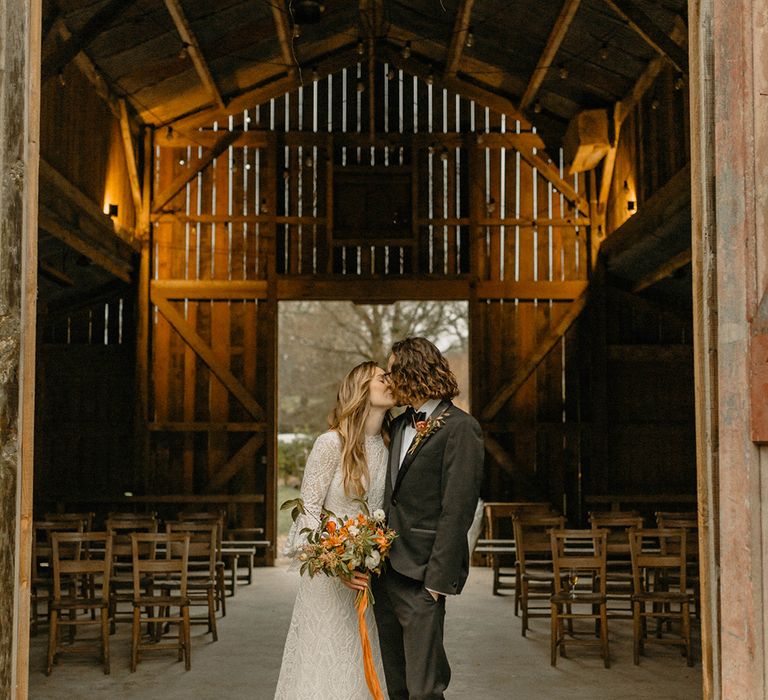 Groom in black tie kisses bride in lace wedding dress with bell sleeves and orange and white bouquet 