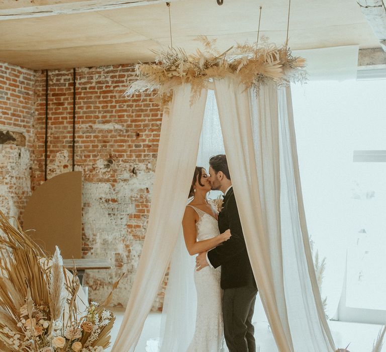 Bride and groom kiss under their white wedding drape altar decor with candles, dried flowers and cream rugs