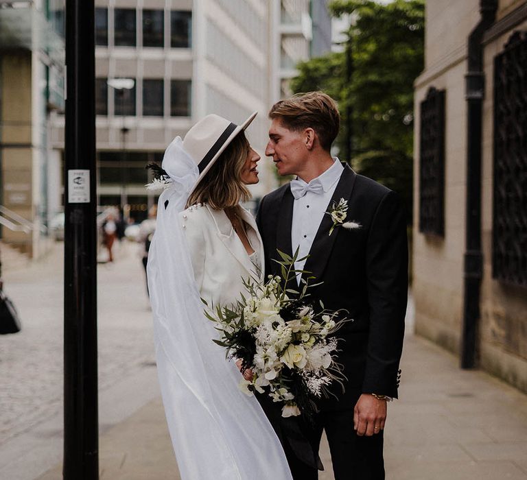 Bride in white trouser Nadine Merabi pearl suit with groom in black tie and pale blue bow tie for town hall wedding 