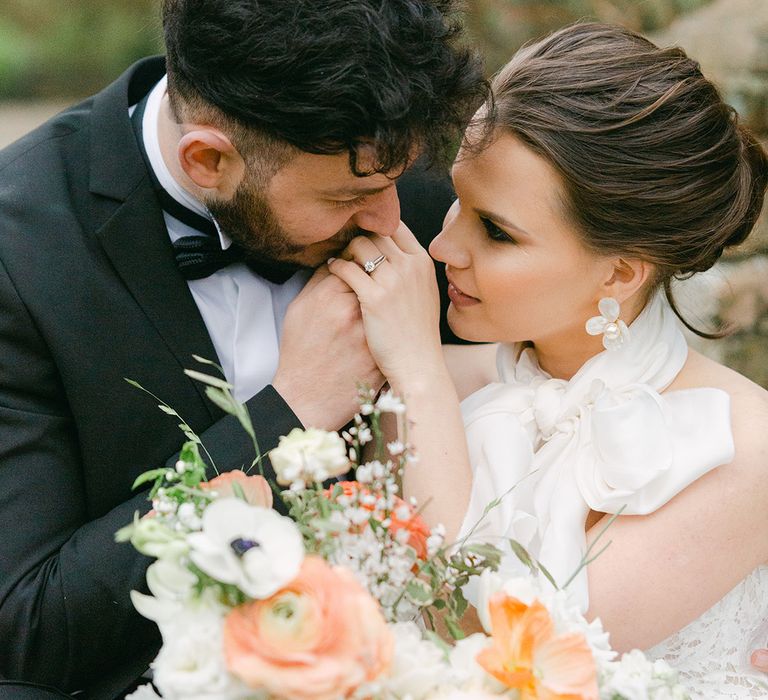 Groom in a tuxedo kissing his brides hands with a chic pinned wedding hairstyle and flower earrings 