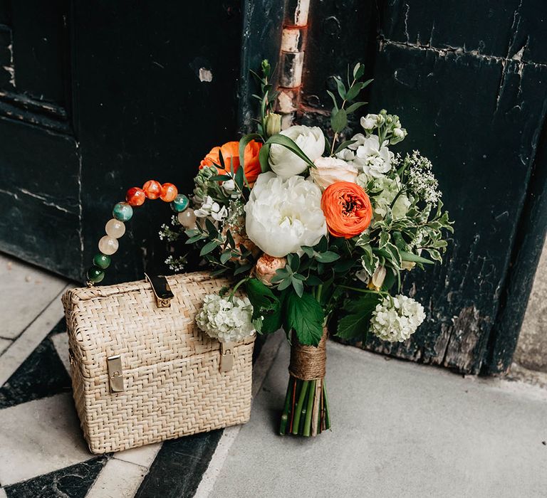 White & orange floral bridal bouquet next to wicker purse complete with brightly beaded handle 