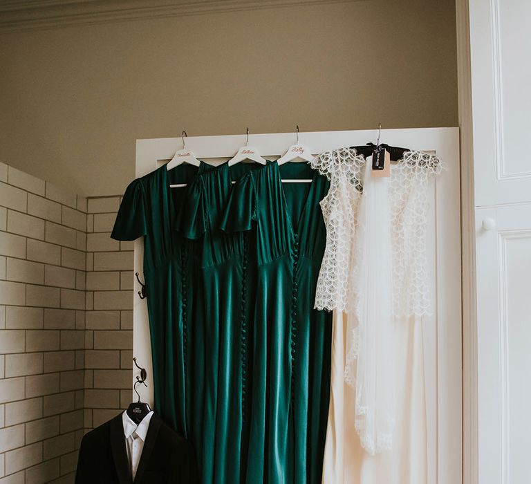 Emerald green satin bridesmaids dresses hang beside Kate Beaumont wedding gown and suit 