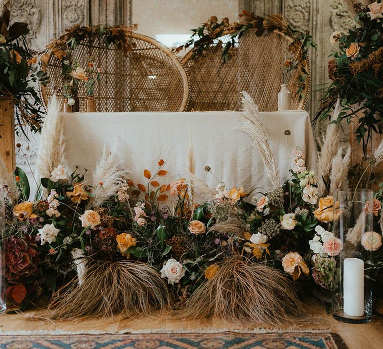 Boho altar setting with peacock chairs, pampas grass, orange wedding flowers and candles in hurricane vases 